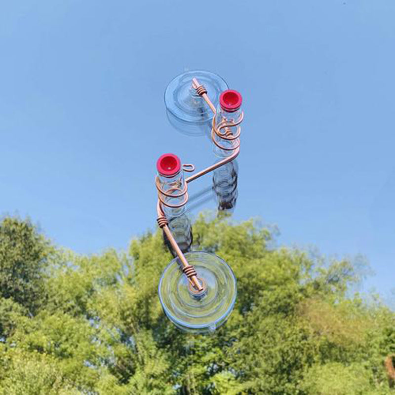 Glass and copper garden bird feeder with two red feeding ports mounted on a window with trees in background