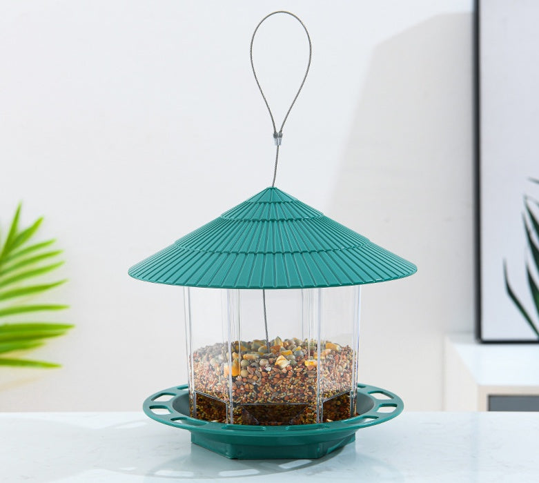 Green roof transparent bird feeder filled with mixed seeds hanging indoors on a white surface